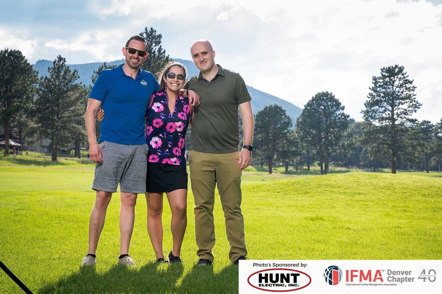 Three people standing on a grassy field with trees and mountains in the background. Two men have their arms around a woman.