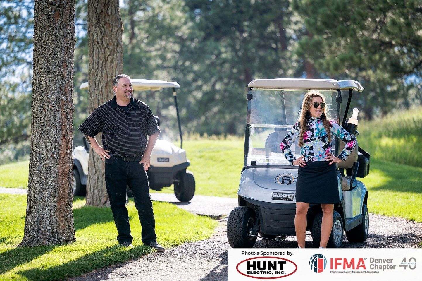 Two people standing by golf carts on a course. Man in black, woman in patterned shirt and skirt.