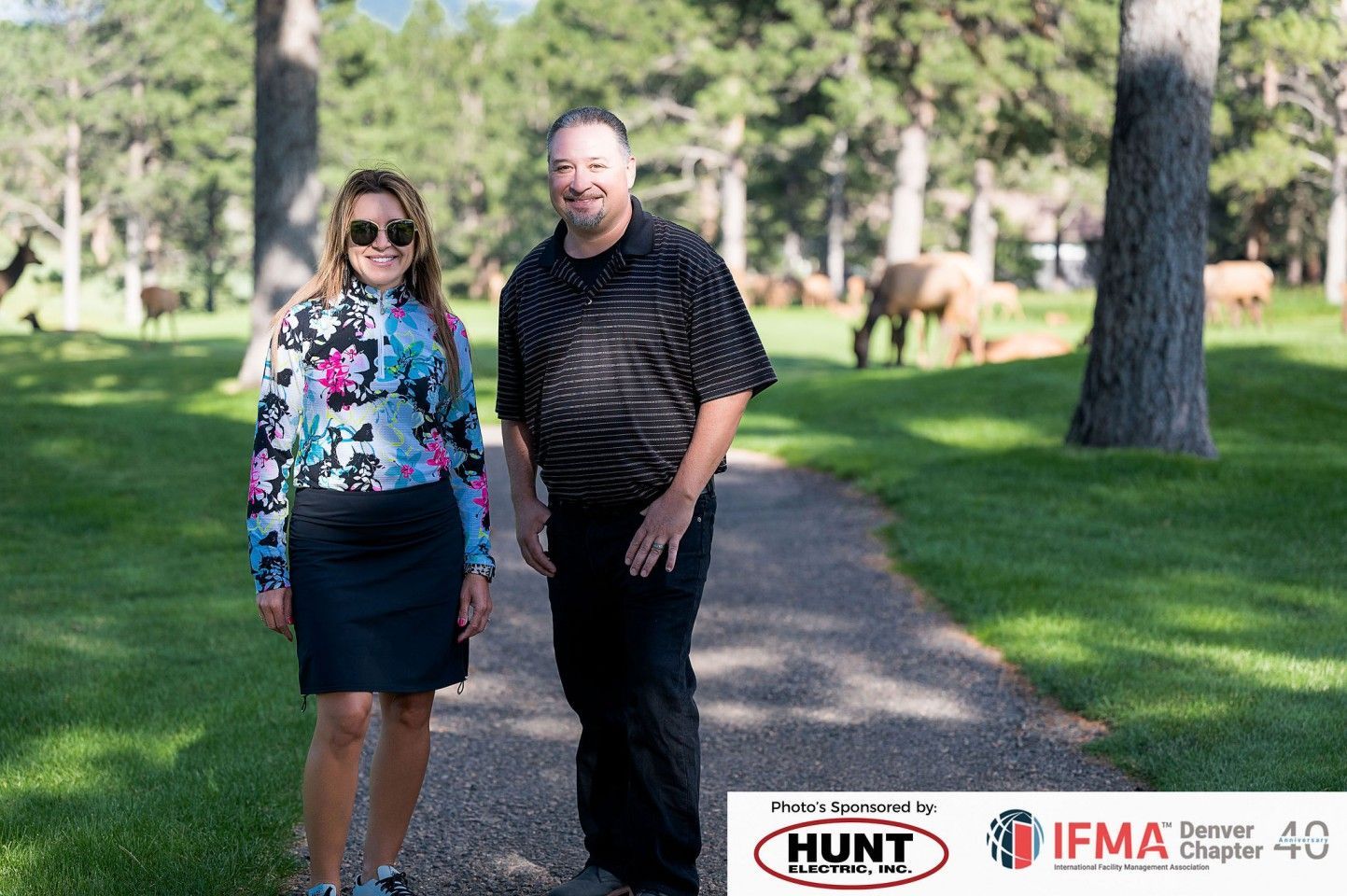 Woman in floral jacket and man in black shirt stand on a path. Deer graze in the background.