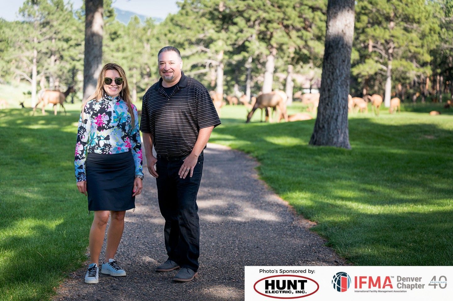A man and woman stand on a path in a park with elk in the background.
