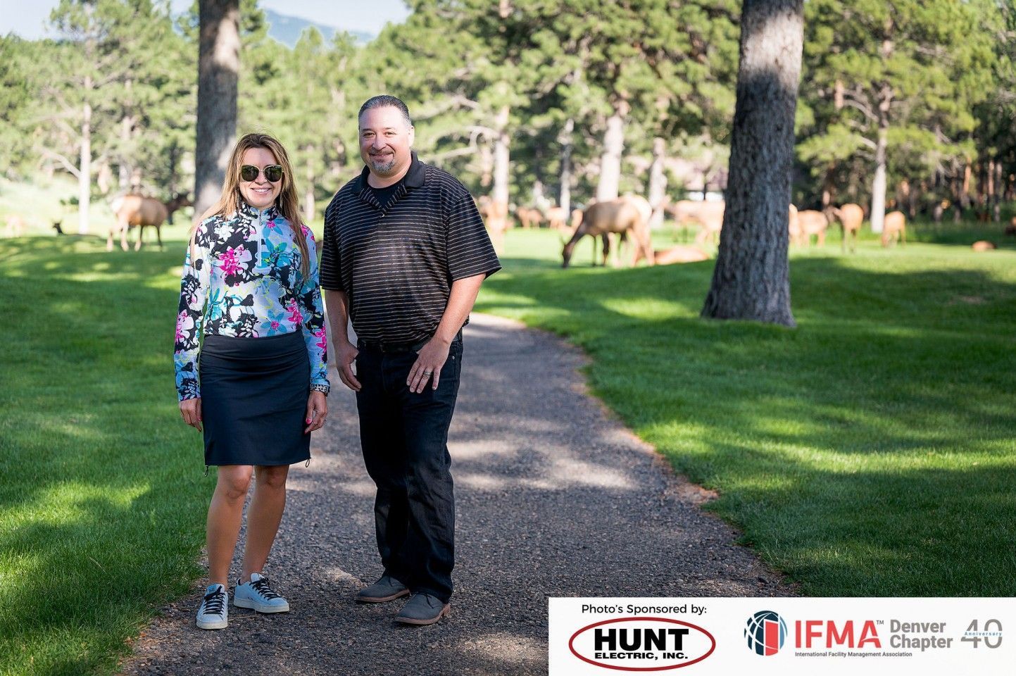 Man and woman stand on a path with elk in the background, trees, and grass.
