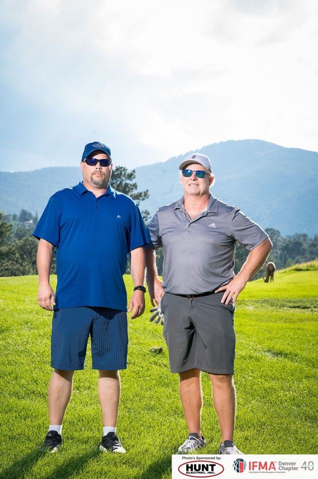 Two men in golf attire on a green course. One in blue, one in gray, with mountains in the background.