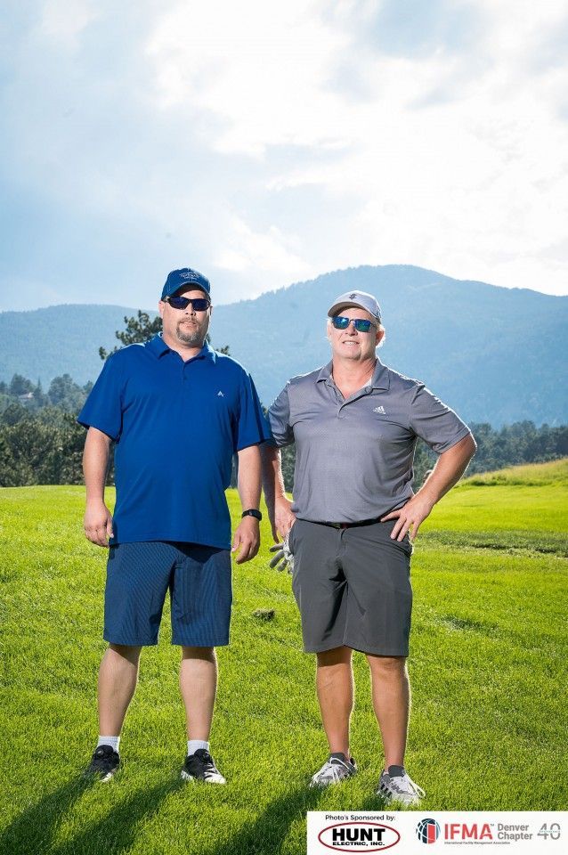 Two men on a golf course posing. One wears blue, the other gray. Mountains in the background.