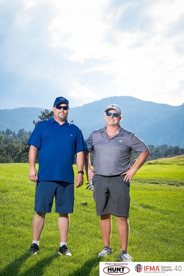 Two men in golf attire stand on a green golf course, mountains in the background, sunny day.