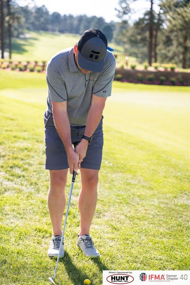 Man preparing to putt a golf ball on a green. Wearing a hat, grey shirt, shorts, and sneakers. Sunny outdoor setting.