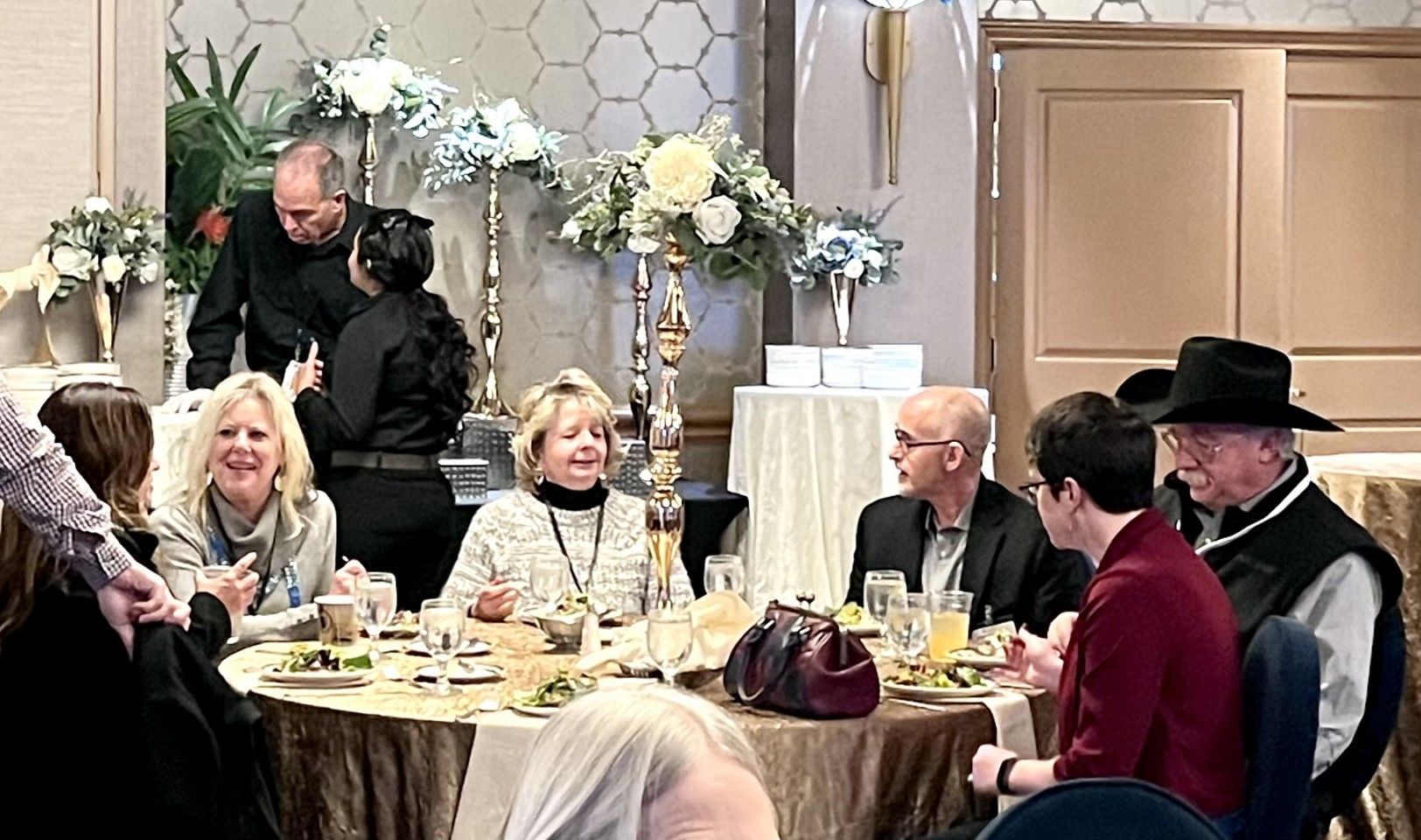 People gathered at a round table, possibly at an event, with centerpieces and food; some are talking and smiling.