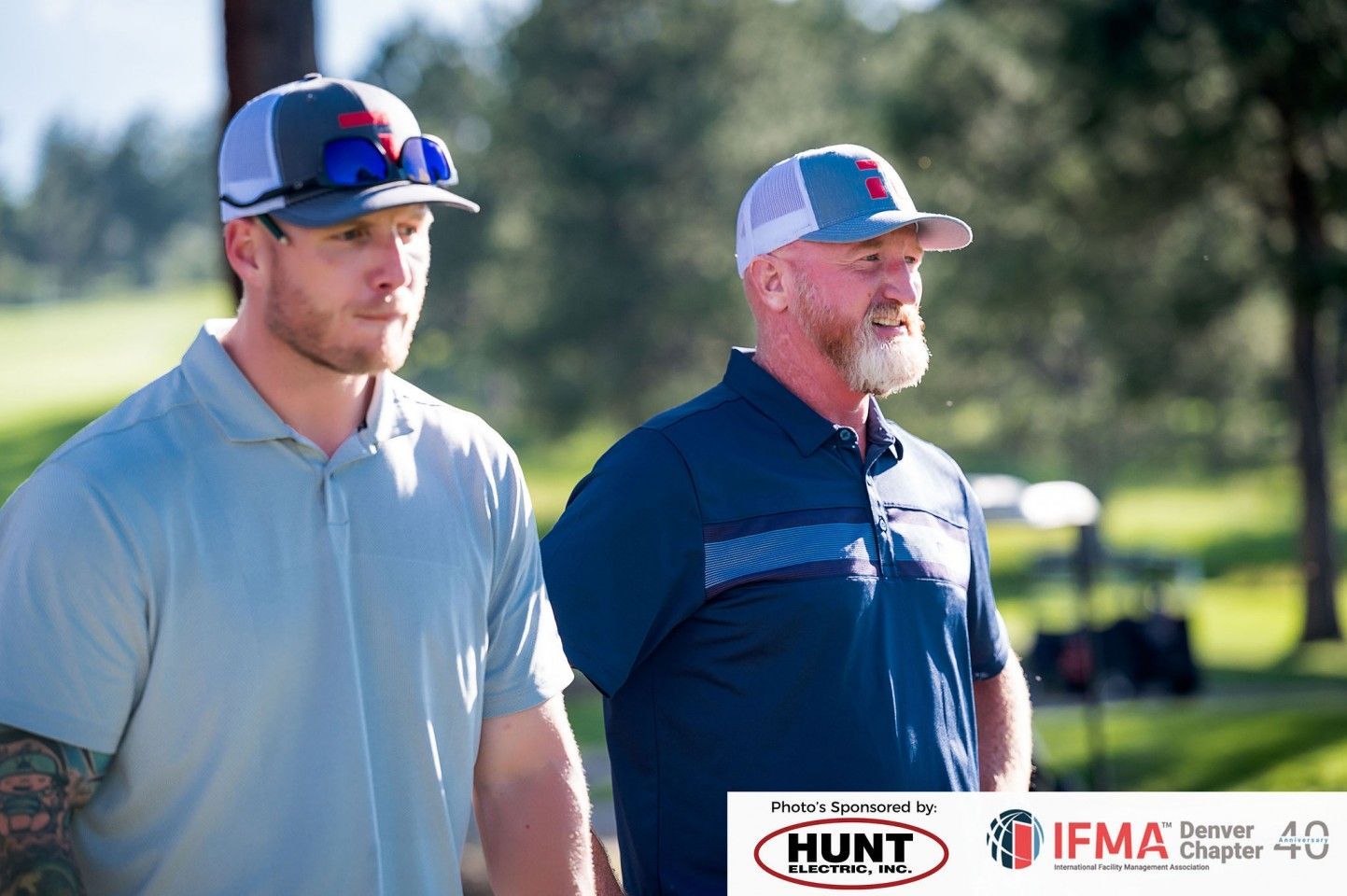 Two men in golf attire on a course. One has a tattoo, the other a beard. They wear caps and look ahead.