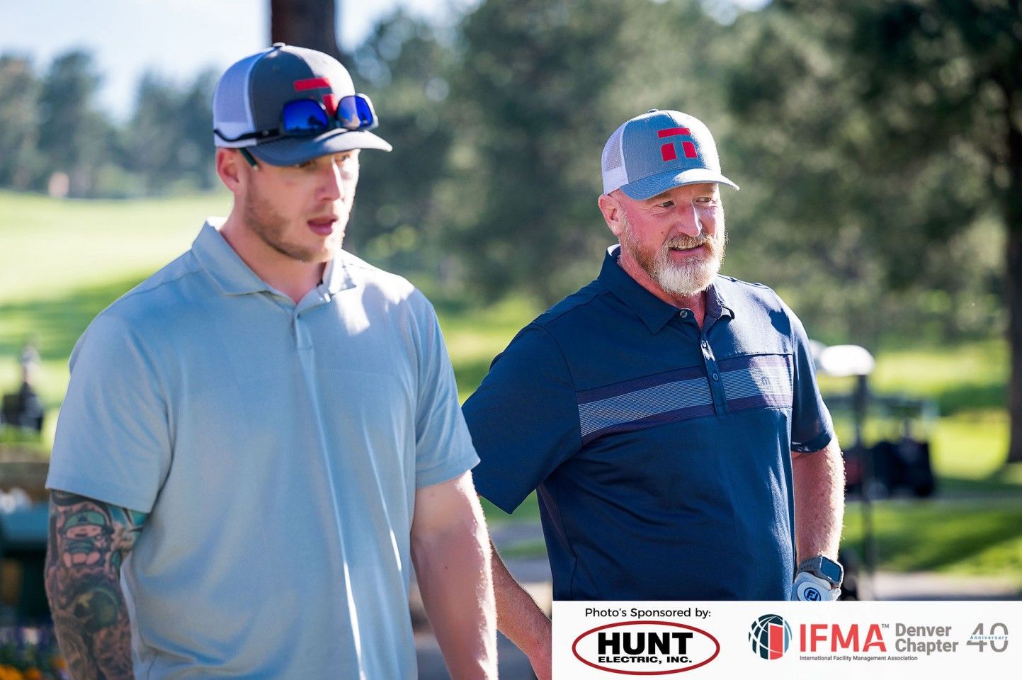 Two men in golf attire walking on a green. One has tattoos and a gray shirt, the other a beard and a navy shirt.