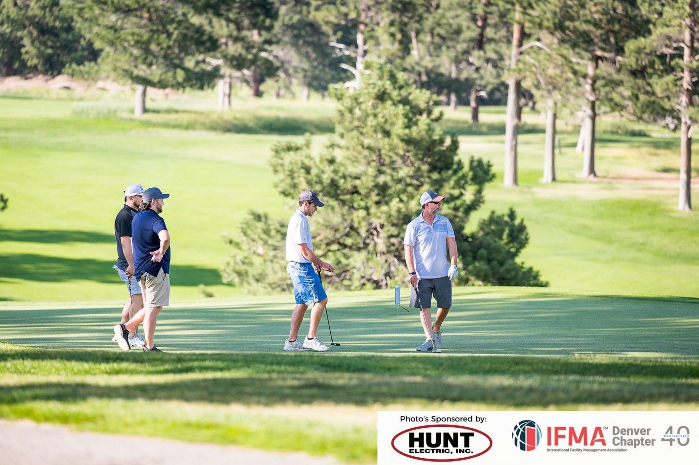 Golfers on a sunny course. Four men walking and preparing to play. Trees and green grass in the background.