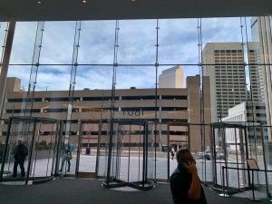 Exterior view through glass walls with revolving doors; buildings and cloudy sky visible.