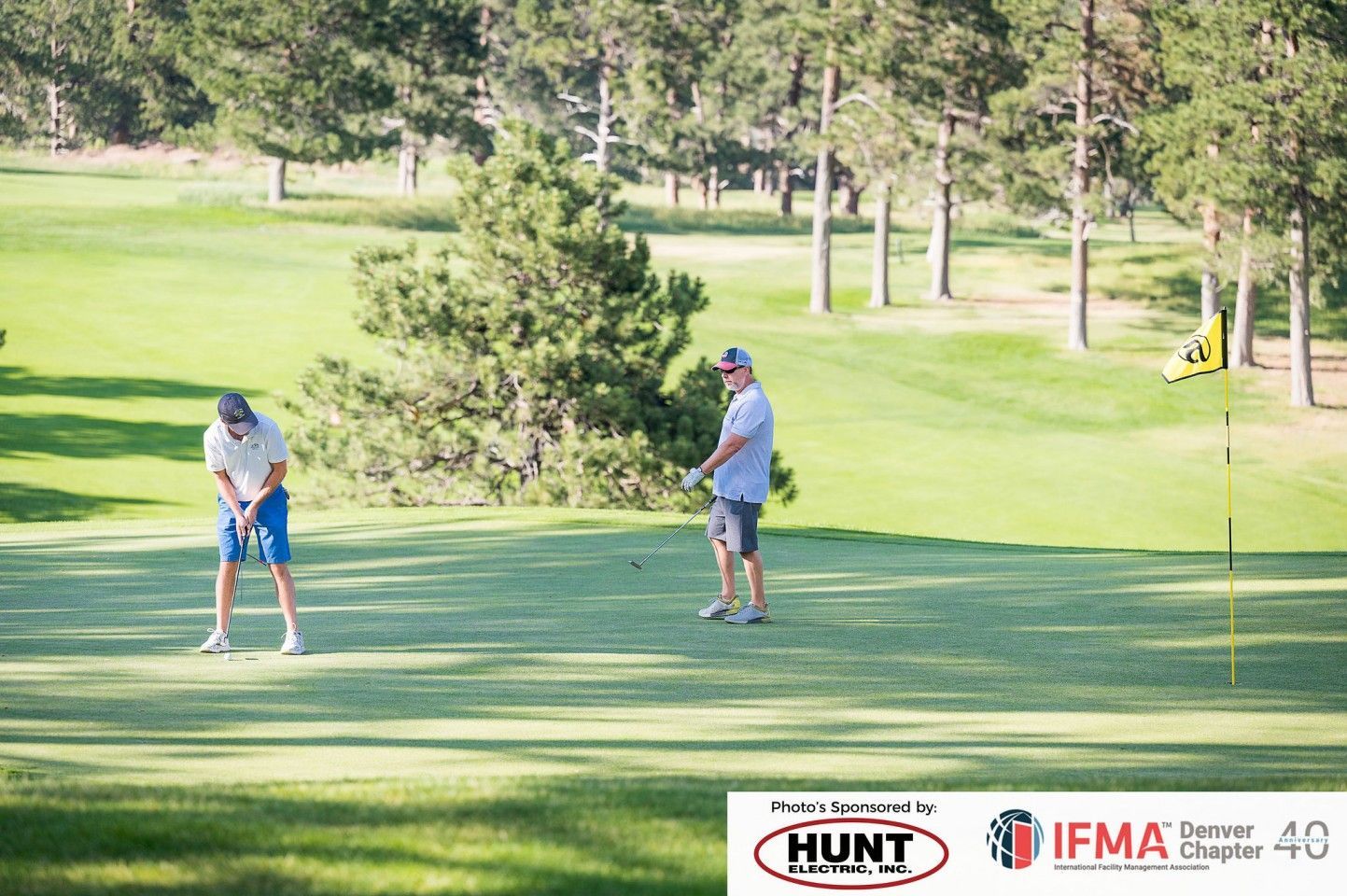 Two golfers on a green putting, sunny day. Trees and flag in the background.