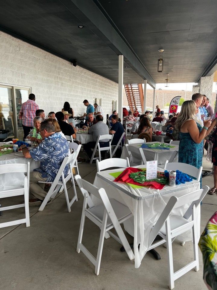 Outdoor event: People seated at tables under a covered patio. White chairs, tables with party favors, and food.