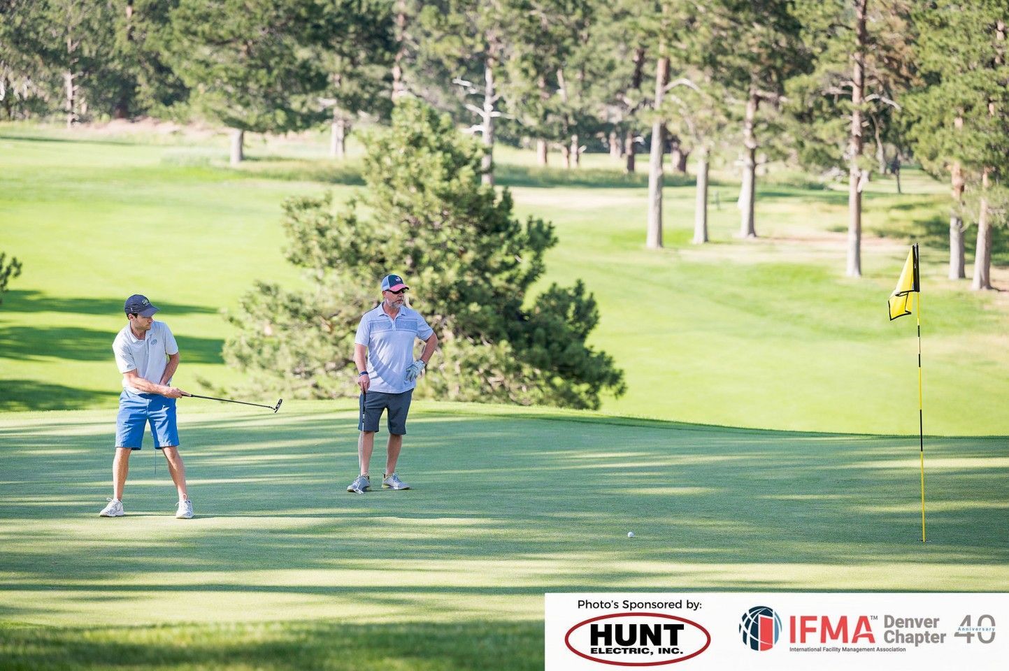 Two men playing golf on a green course. One swings a club, the other watches.