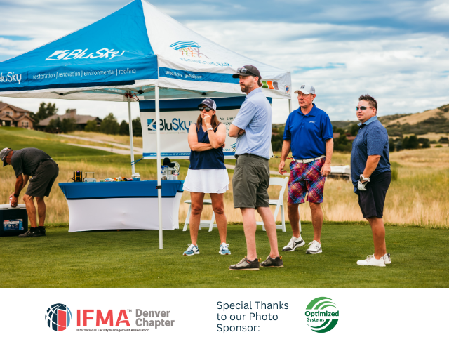 People at a golf event under a branded tent on a green field. Some are talking, wearing golf attire.