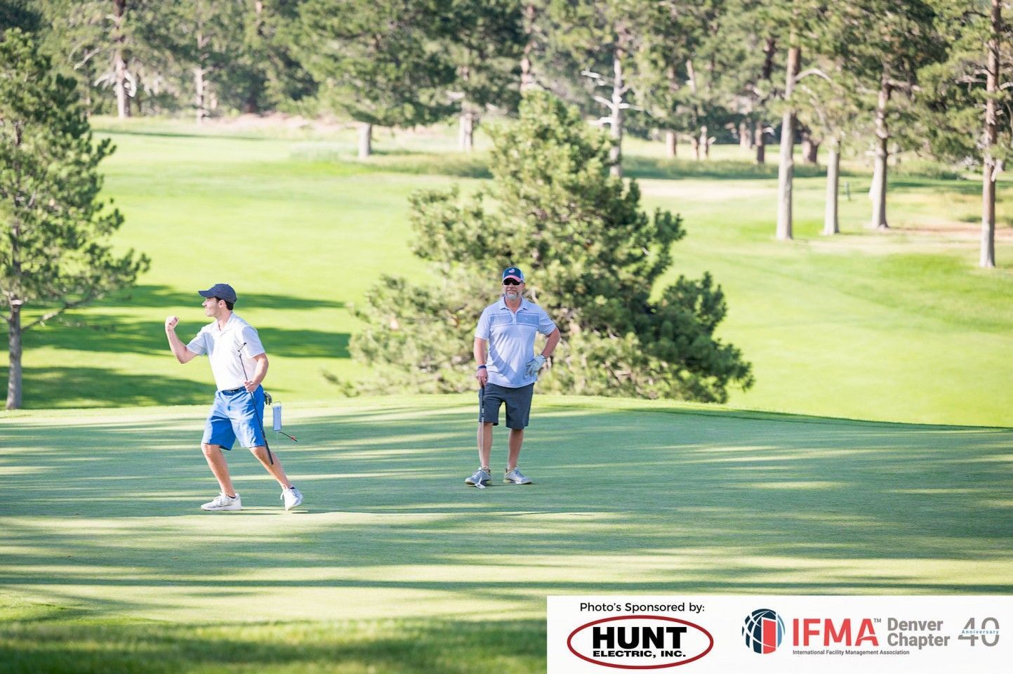 Two men on a sunny golf course. One gestures with a golf club while the other watches.
