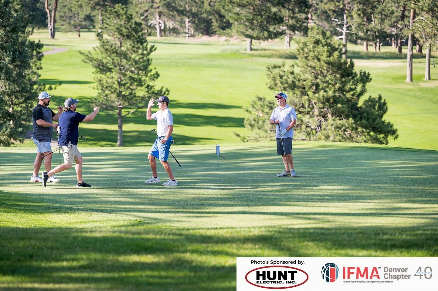 Four men on a golf course: two high-fiving, two watching, sunny day.