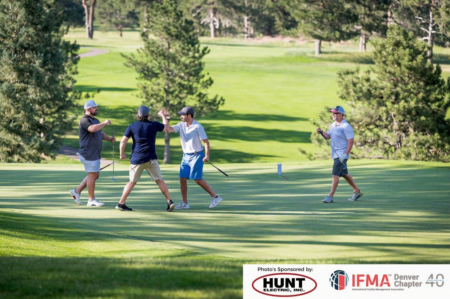 Four golfers on a green celebrate. One high-fives another, others smile near a flag on a sunny course.
