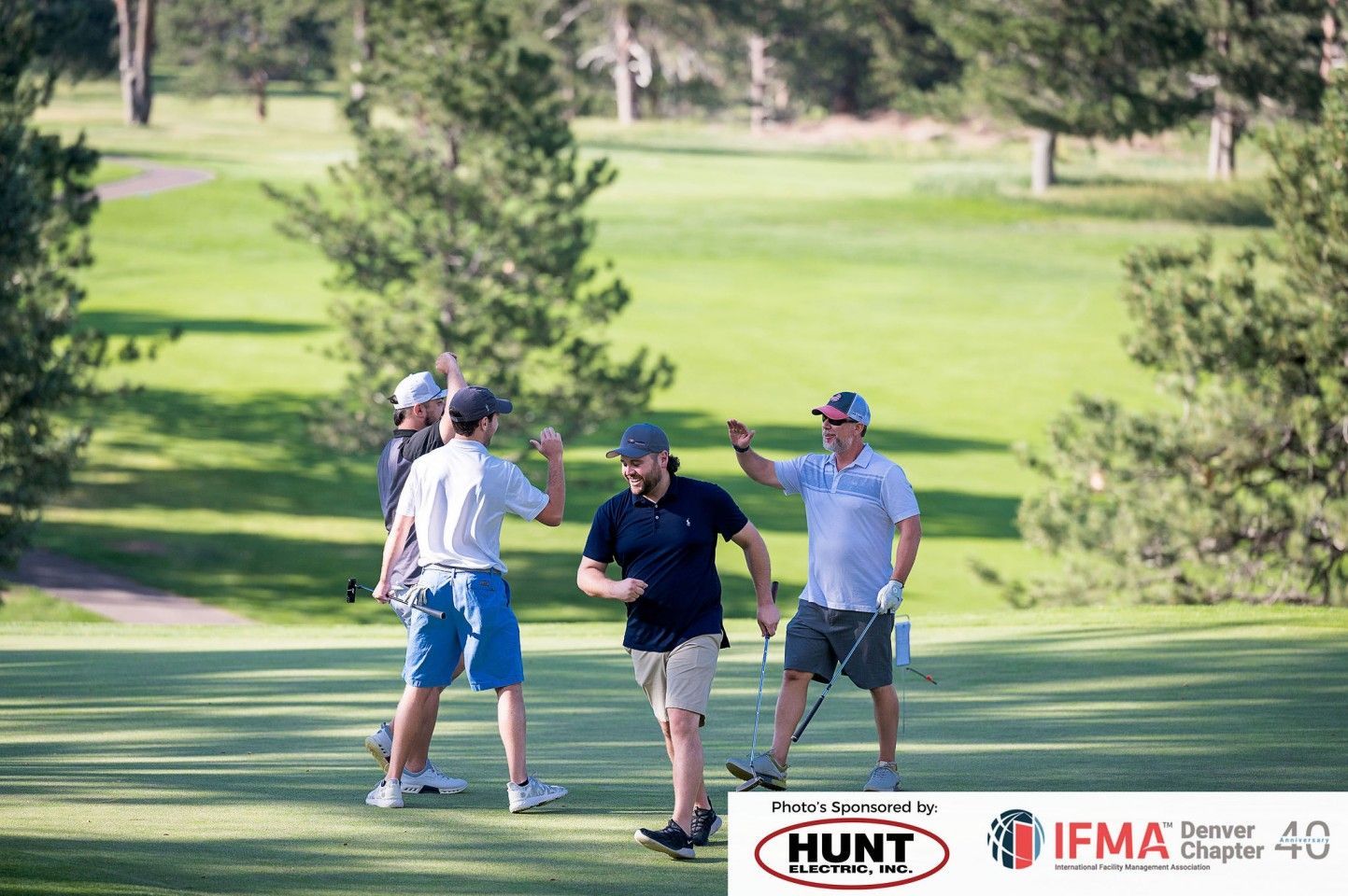 Four golfers celebrate on a green course, high-fiving. Sunny day, trees in background, IFMA banner.