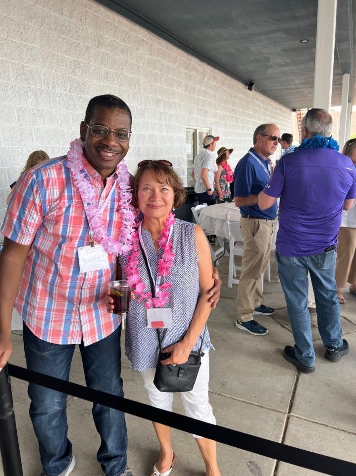 Two people in leis smile, posing for a photo. They are outside near a fence.