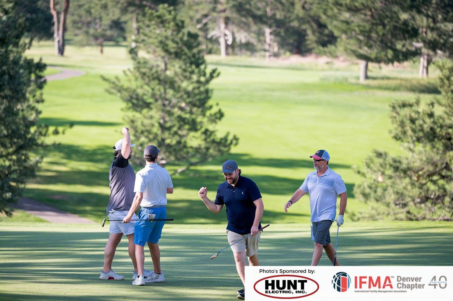 Four golfers on a golf course celebrating, one man pumping fist. Green grass, trees, sunny day.