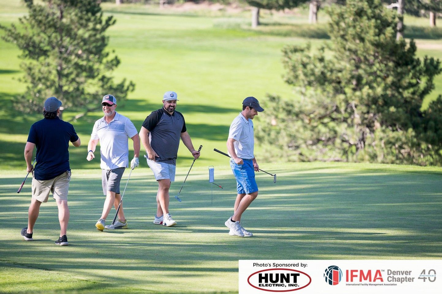 Four people playing golf on a green course on a sunny day. They are holding golf clubs.