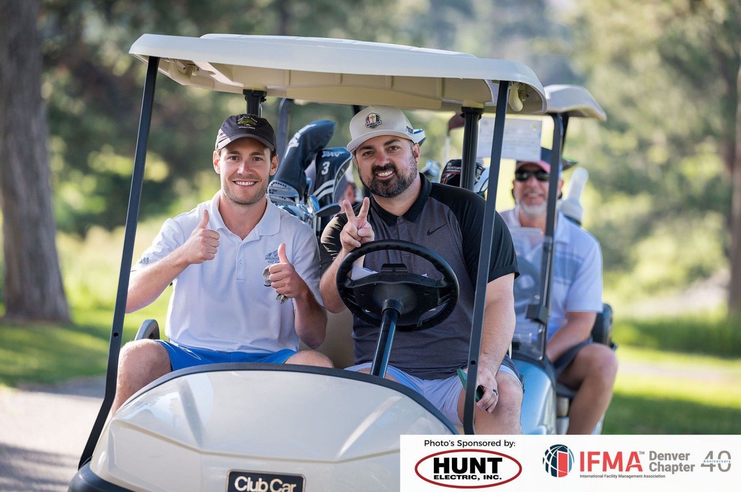 Three men in a golf cart; two smiling, giving thumbs-up and peace signs.  Green fairway background, sunny.