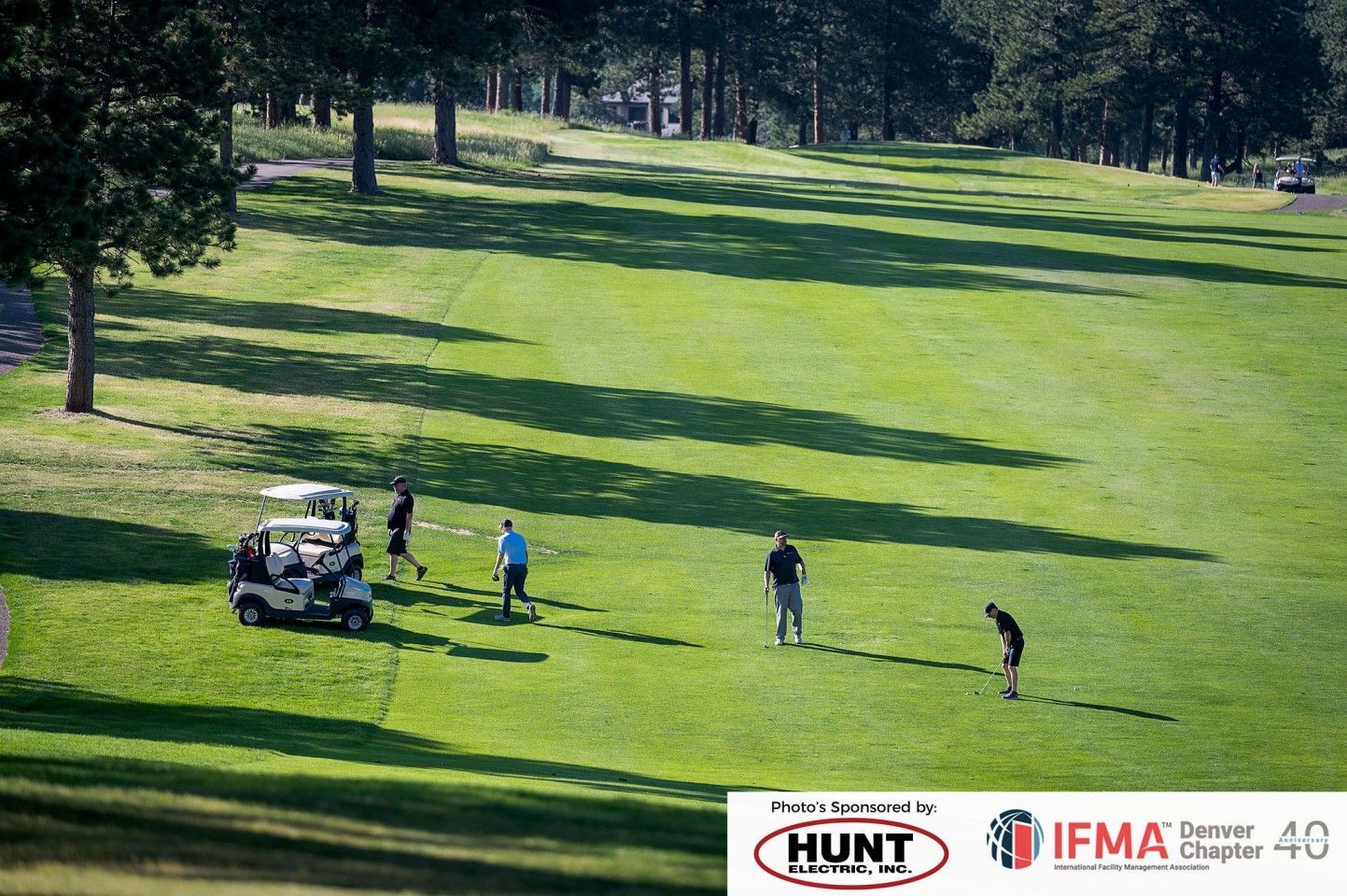 Golfers on a sunny fairway; two golf carts. Green grass, trees, blue sky.