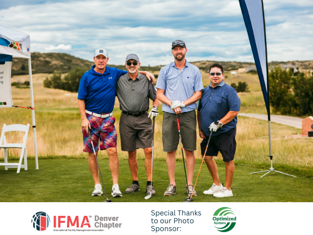 Four men pose on a golf course. They are holding golf clubs and smiling. There is a tent and flag in the background.
