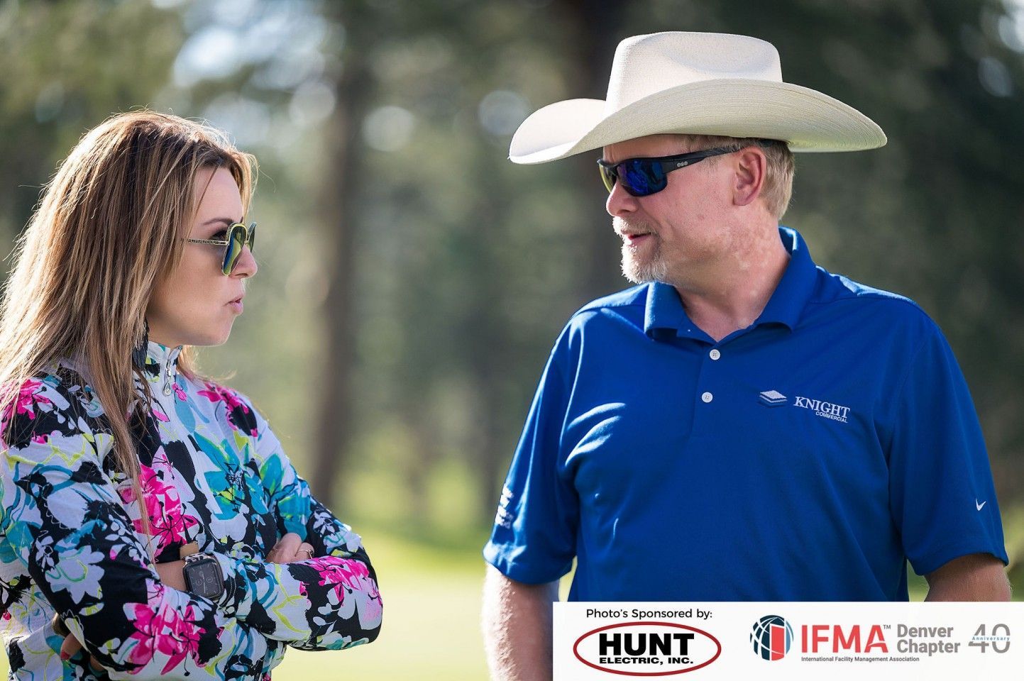 Woman and man in sunglasses conversing outdoors. Man wears cowboy hat and blue shirt.