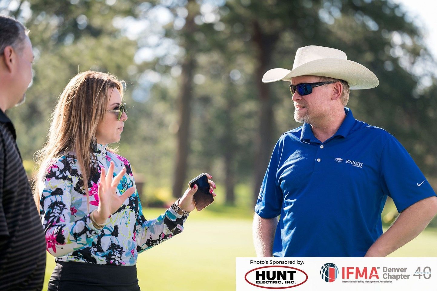 Woman talking to two men on a golf course; one man in cowboy hat.