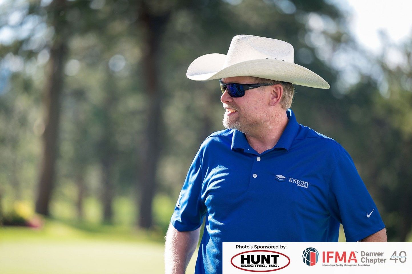 Man in cowboy hat and sunglasses on a golf course, wearing a blue polo shirt, smiling.