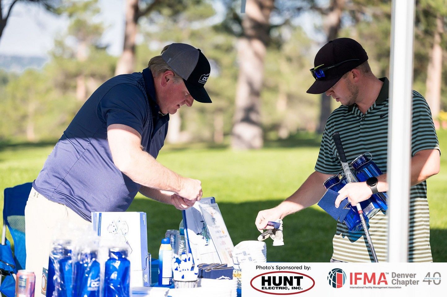 Two men at an outdoor table with items. One in a baseball cap is reaching, the other holds a golf club.
