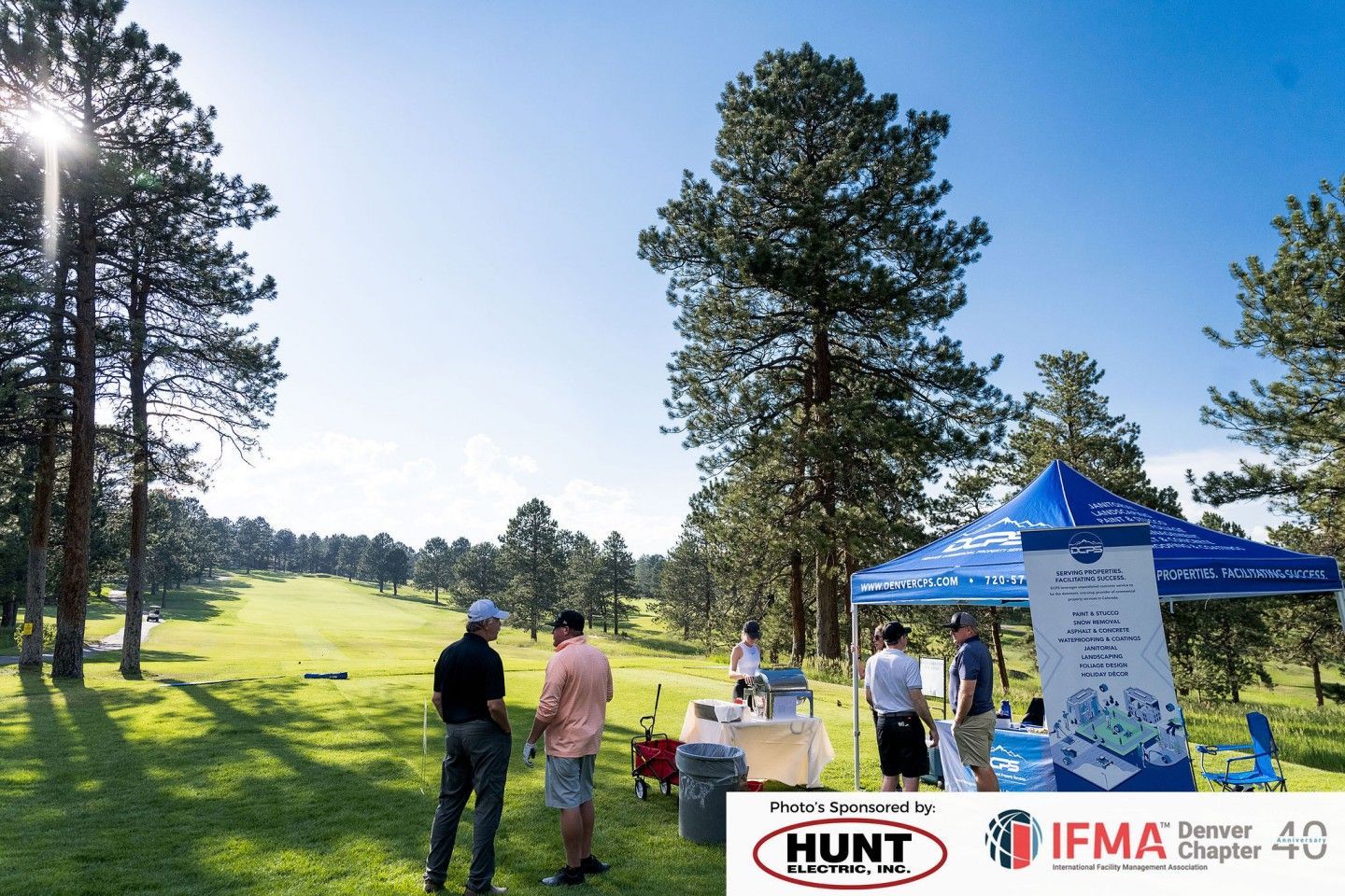 Golf event with people near a tent on a green field. Blue sky, tall trees, sunny day.
