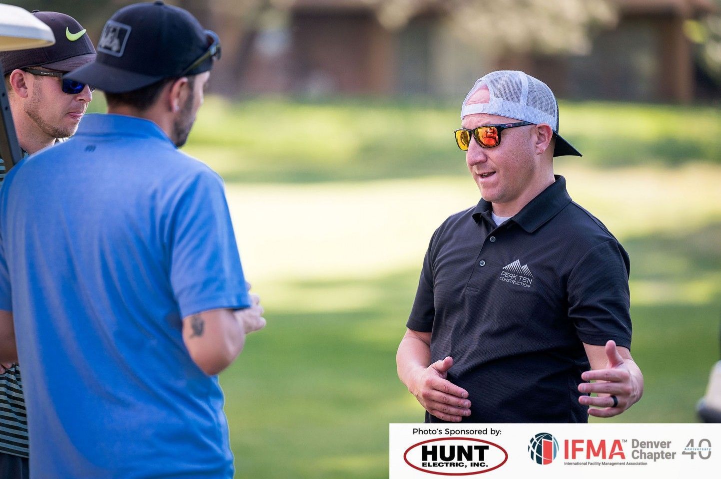 Three men talking outdoors, one in a black polo and mirrored sunglasses gesturing. Green grass and trees in background.
