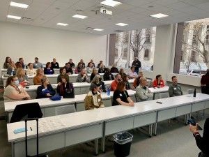People seated in a classroom listening to a presentation. Bright room with large windows.