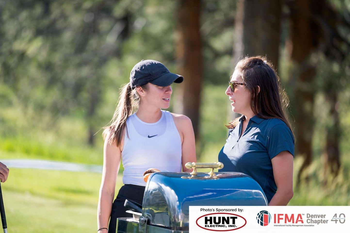 Two women talking near a golf cart, outdoors. One wears a hat, the other sunglasses. Green grass and trees in background.