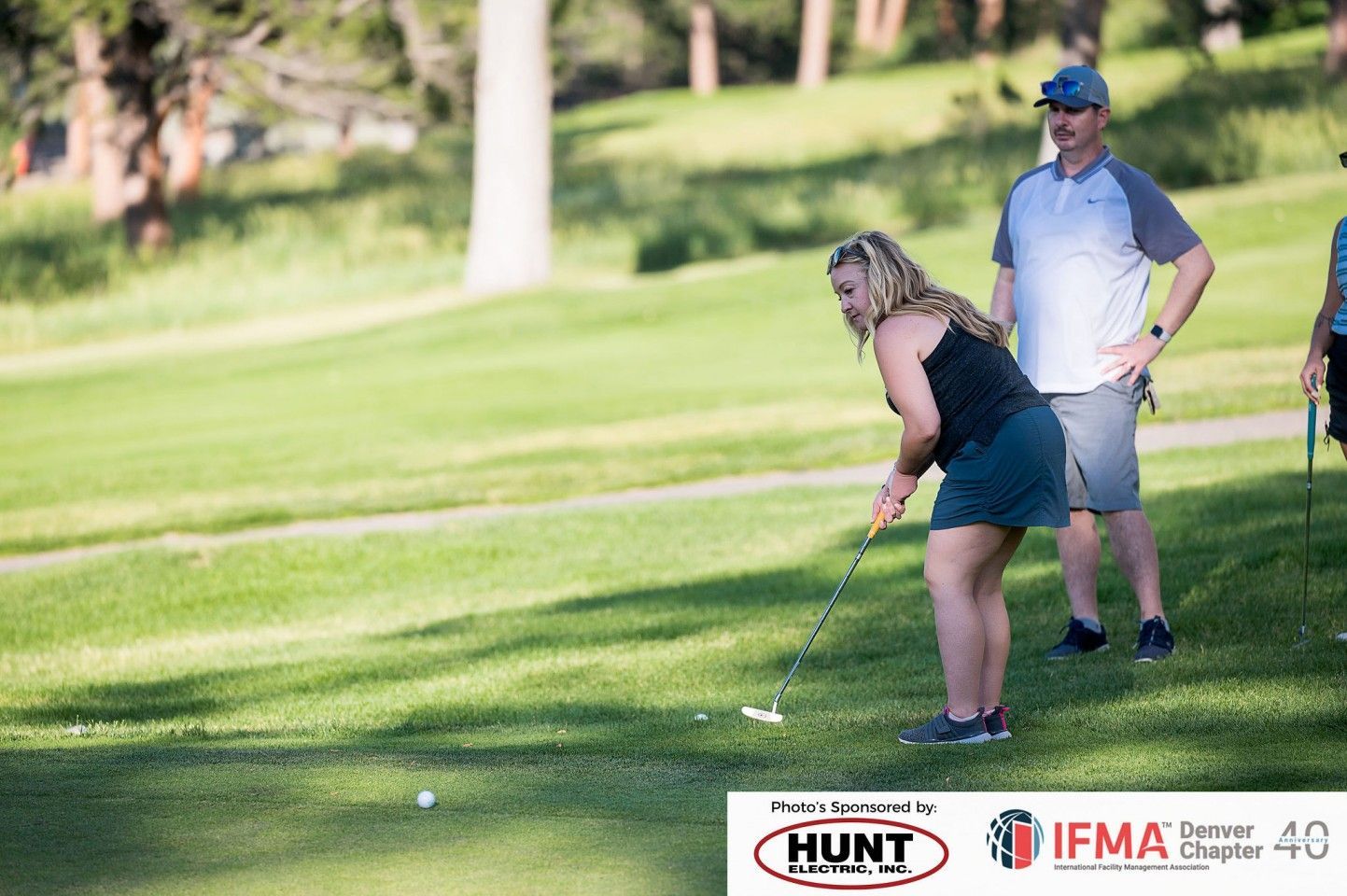 Woman golfing, putting on a green; two people watching in the background. Sunny outdoor setting.