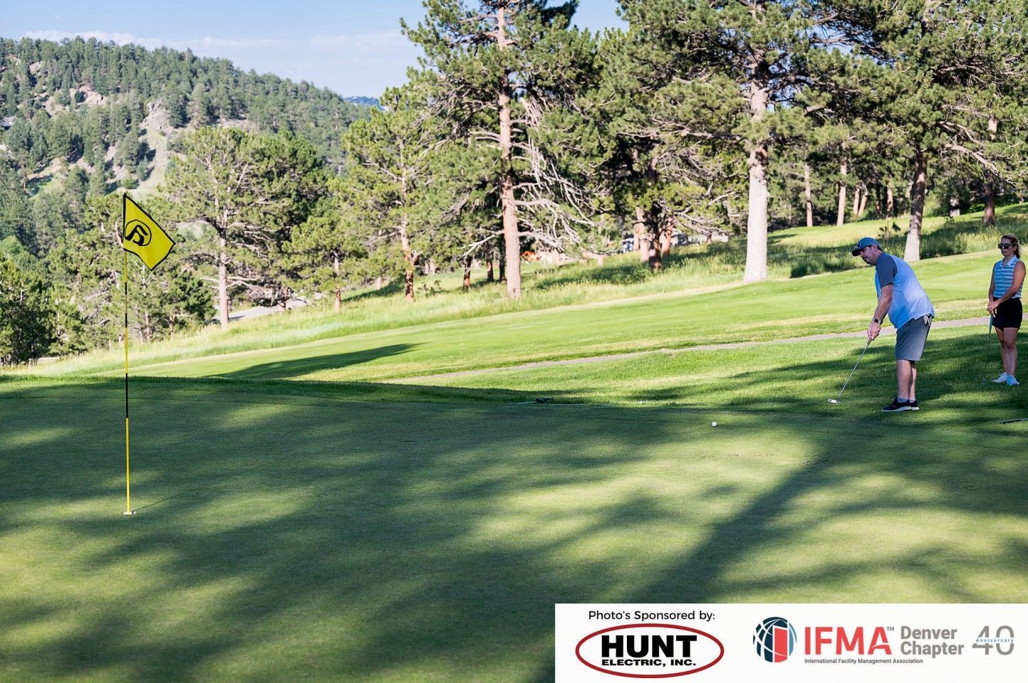 Man putting golf ball on a green, with flag and woman watching; trees and hillside in the background.