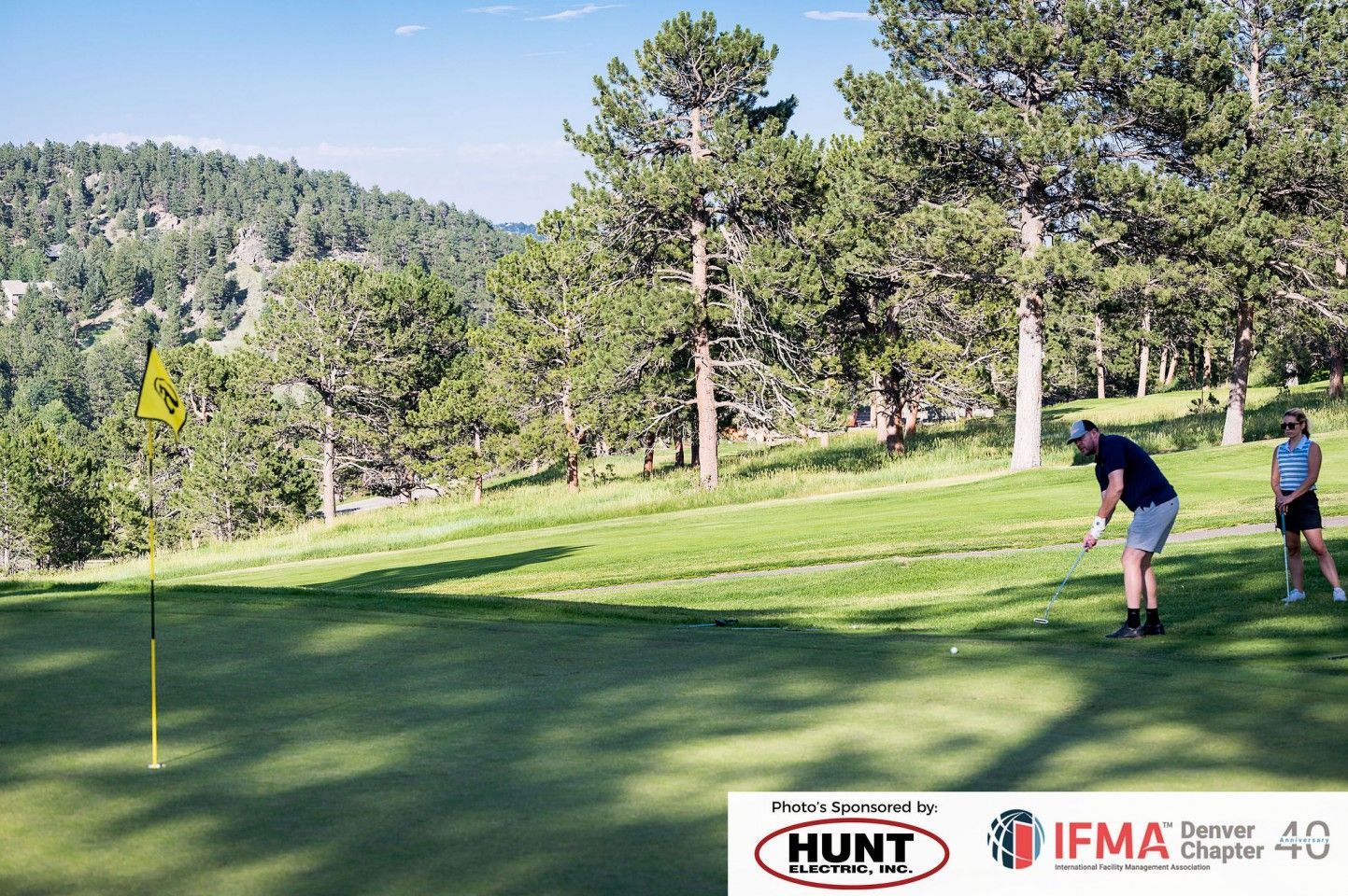 Man golfing, near a flag, with another person on a green hillside. Trees and mountains in background.