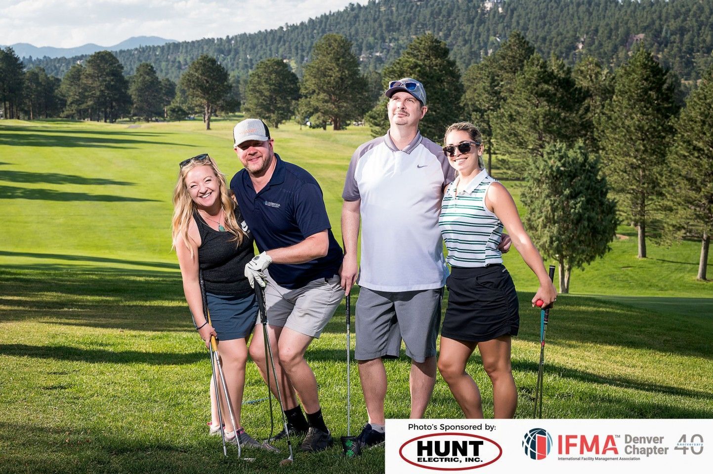 Four golfers pose with clubs on a green. Smiling, against a backdrop of trees and a golf course.