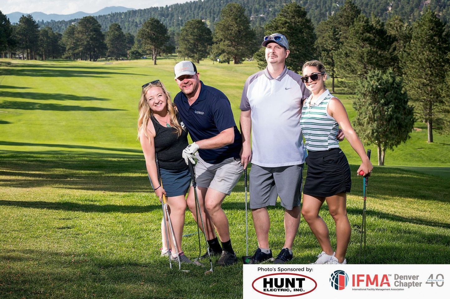 Four people smiling on a golf course, posing with golf clubs. Trees and mountains in the background.
