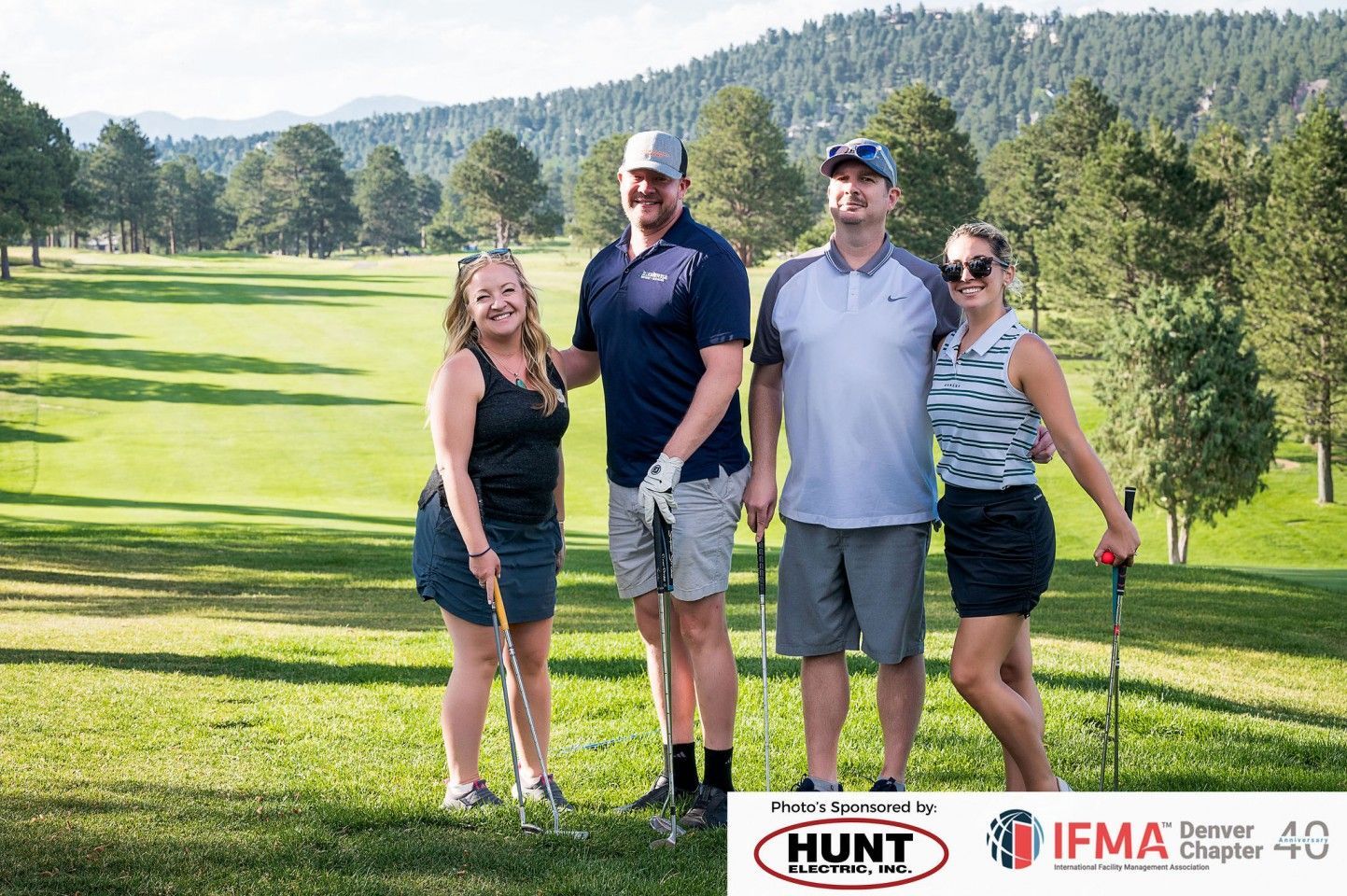Four people smiling on a sunny golf course, holding clubs. Mountains and trees in the background.