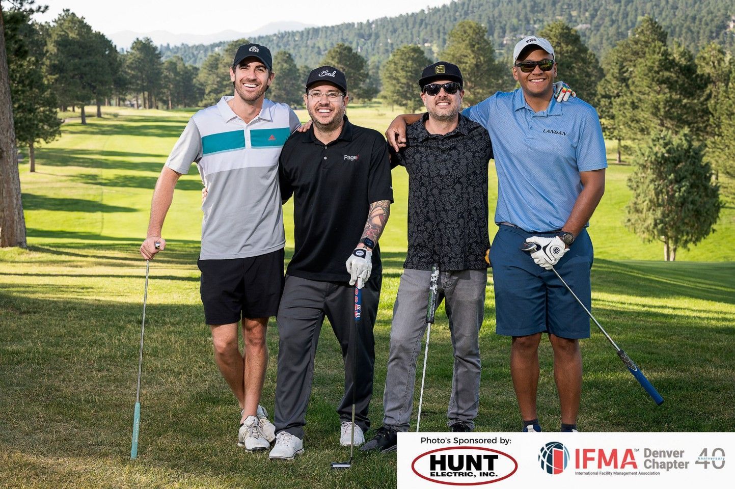 Four men on a golf course pose with clubs. They wear casual golf attire, smiling, with a mountain backdrop.