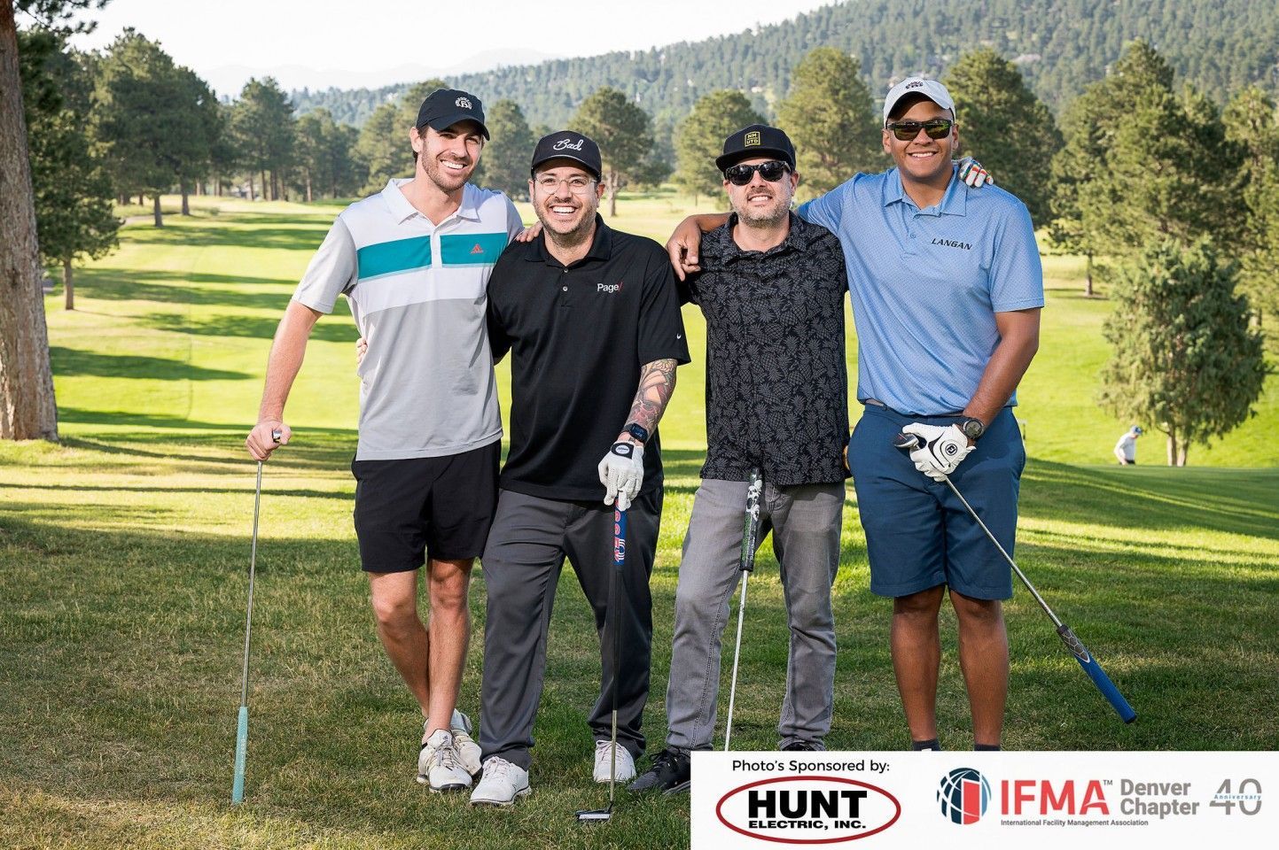 Four men posing together on a golf course, holding clubs. They are smiling, with trees and mountains in the background.