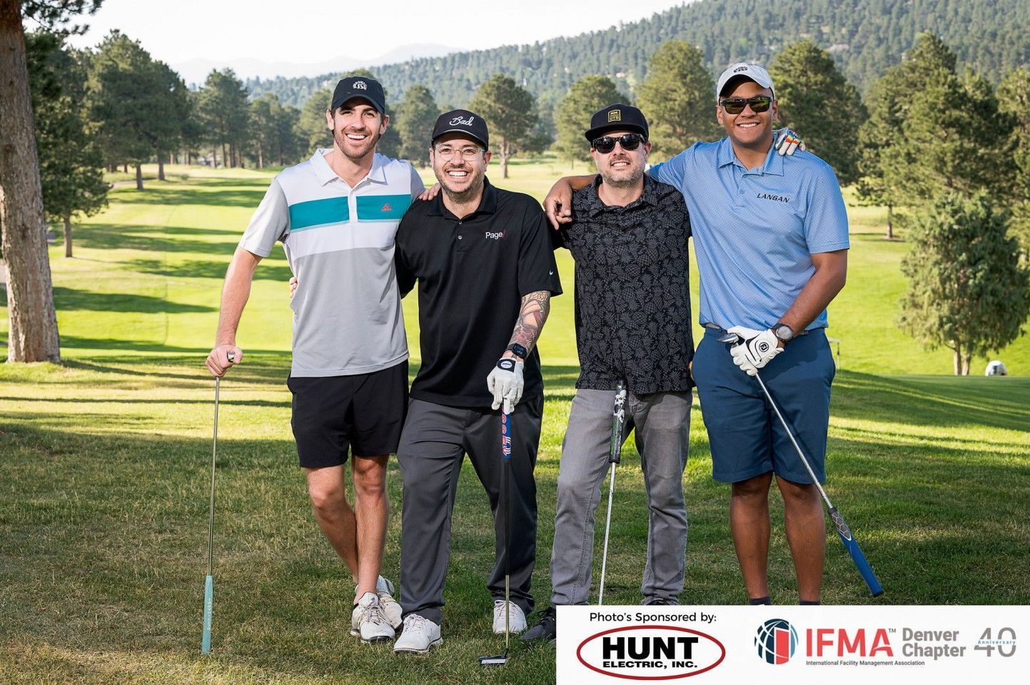Four men on a golf course pose with clubs; green grass, trees, and mountains in the background.