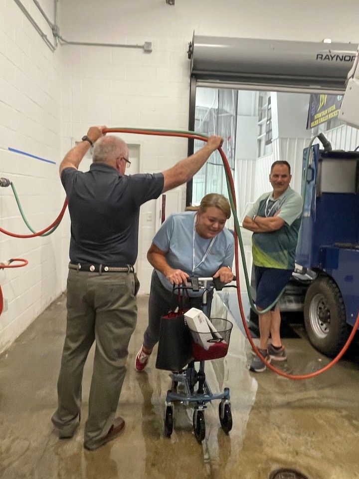 Man holds hose above woman on scooter; another man watches in a room with a truck.