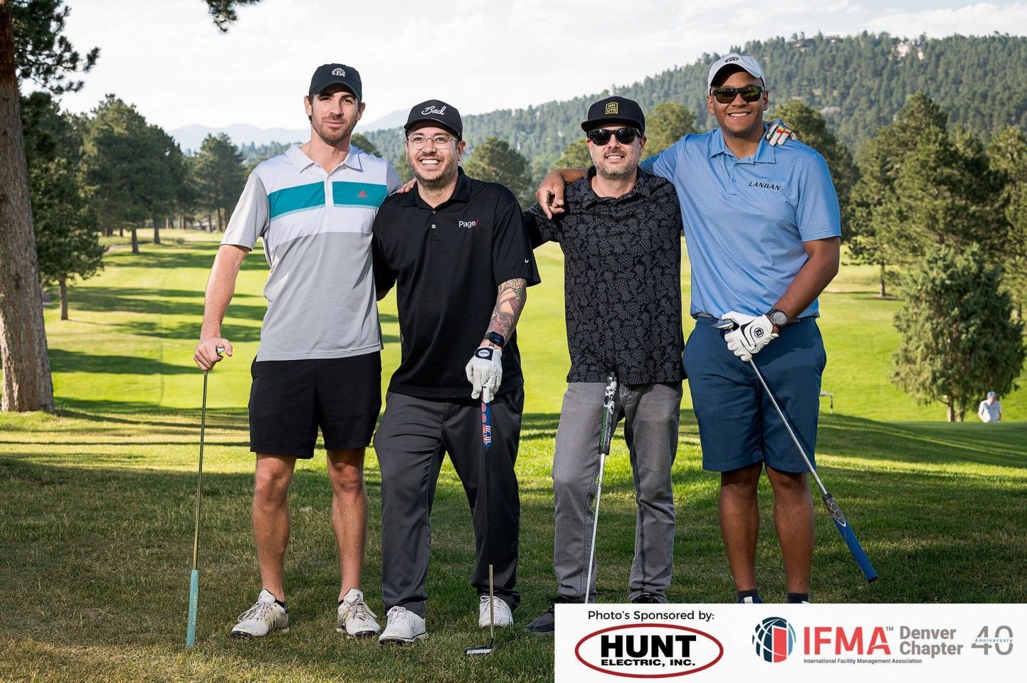Four men on a golf course pose with golf clubs. Trees and mountains in the background. Sponsors' logos at the bottom.