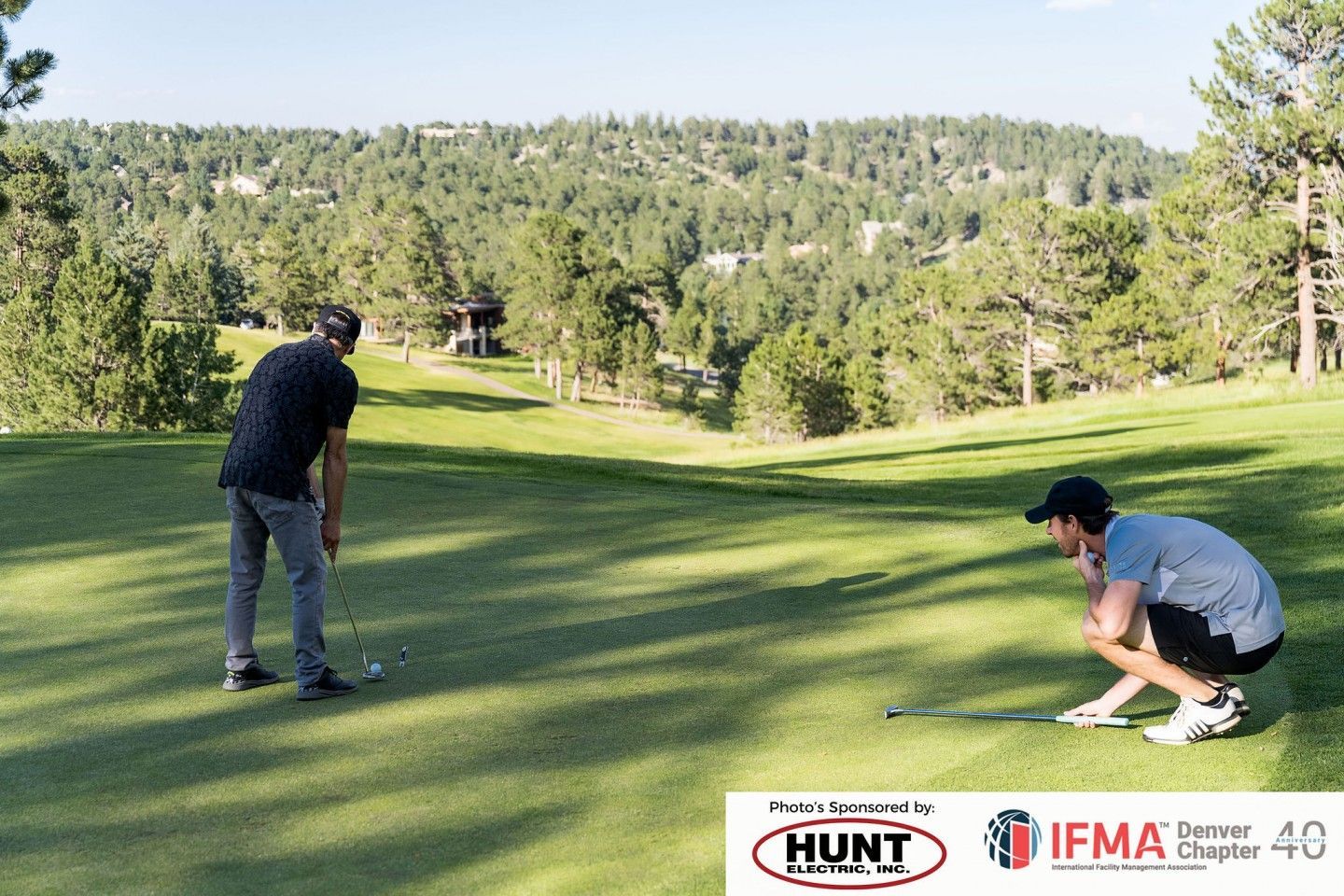 Two golfers on a green: one puts, the other observes. Trees and hills in the background on a sunny day.