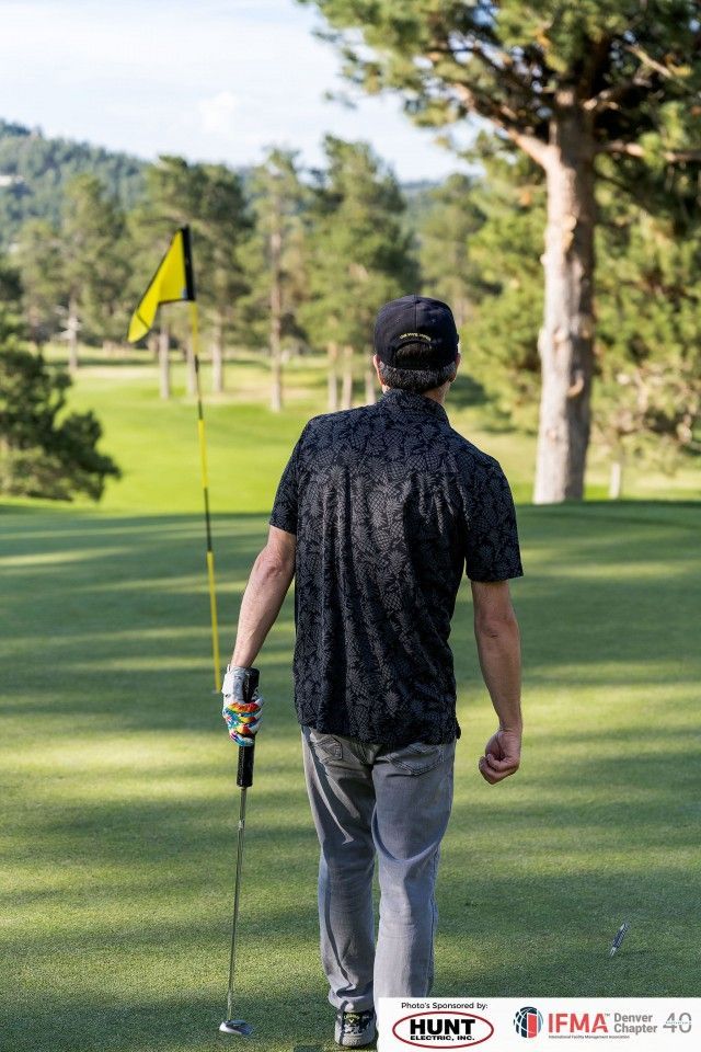Golfer walks on a green toward the flag, holding his club. He wears a dark patterned shirt and gray pants.
