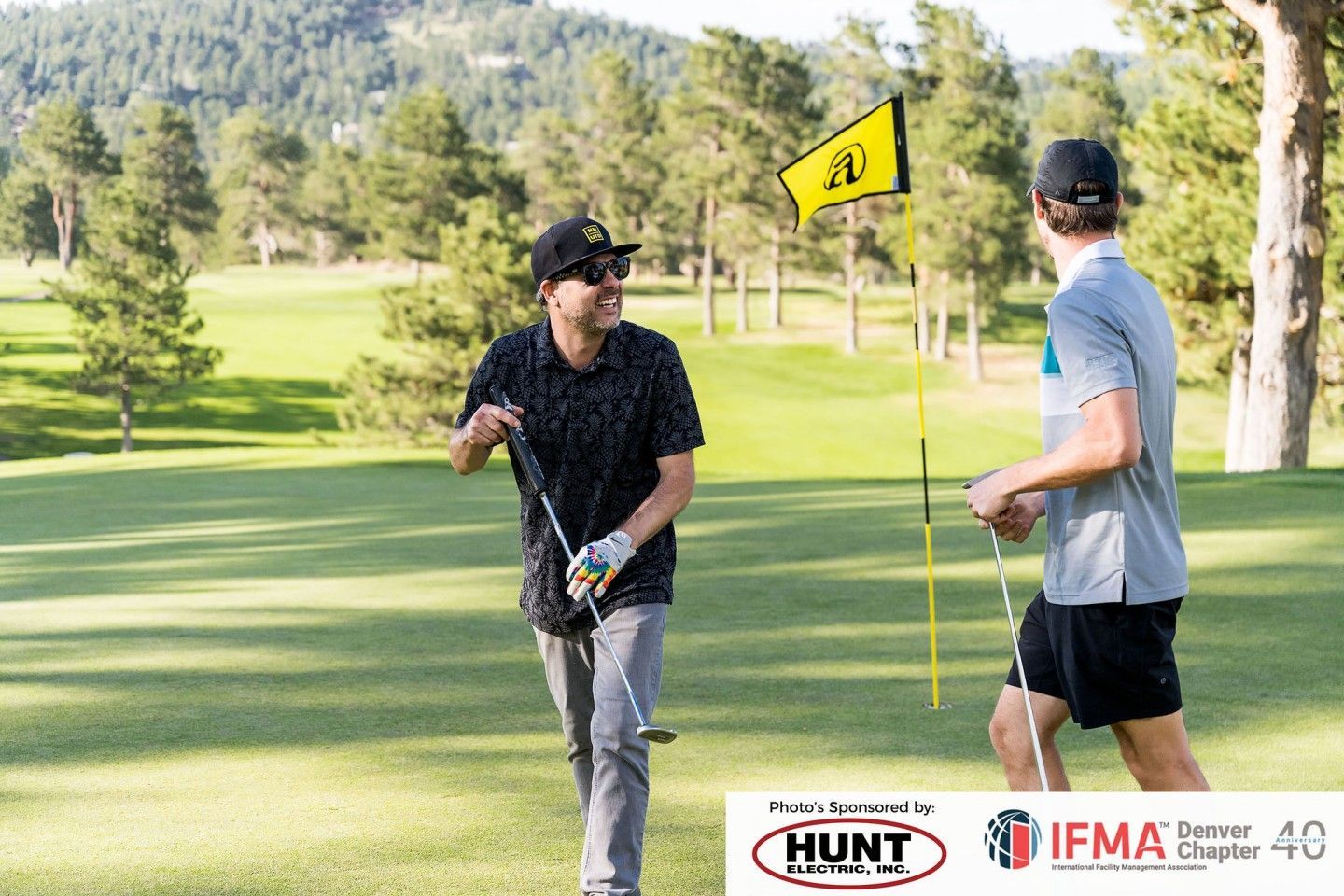 Two men on a golf course. One smiles, holding a club, the other near a flag. Sunny day, green grass.